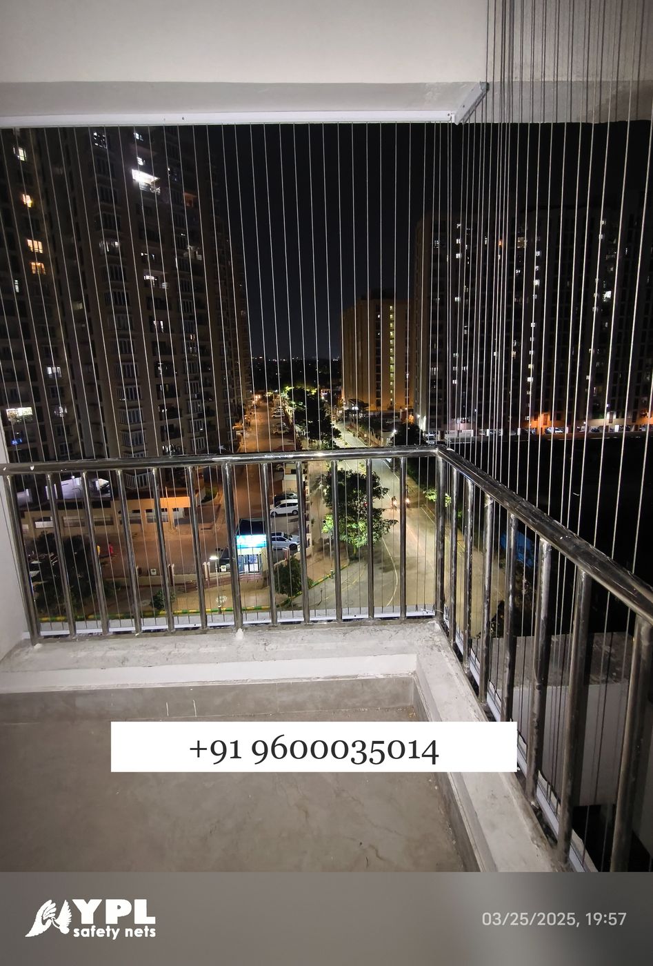 Corner balcony enclosed with vertical invisible grill wires above metal railings, overlooking illuminated apartment towers and streets at night.