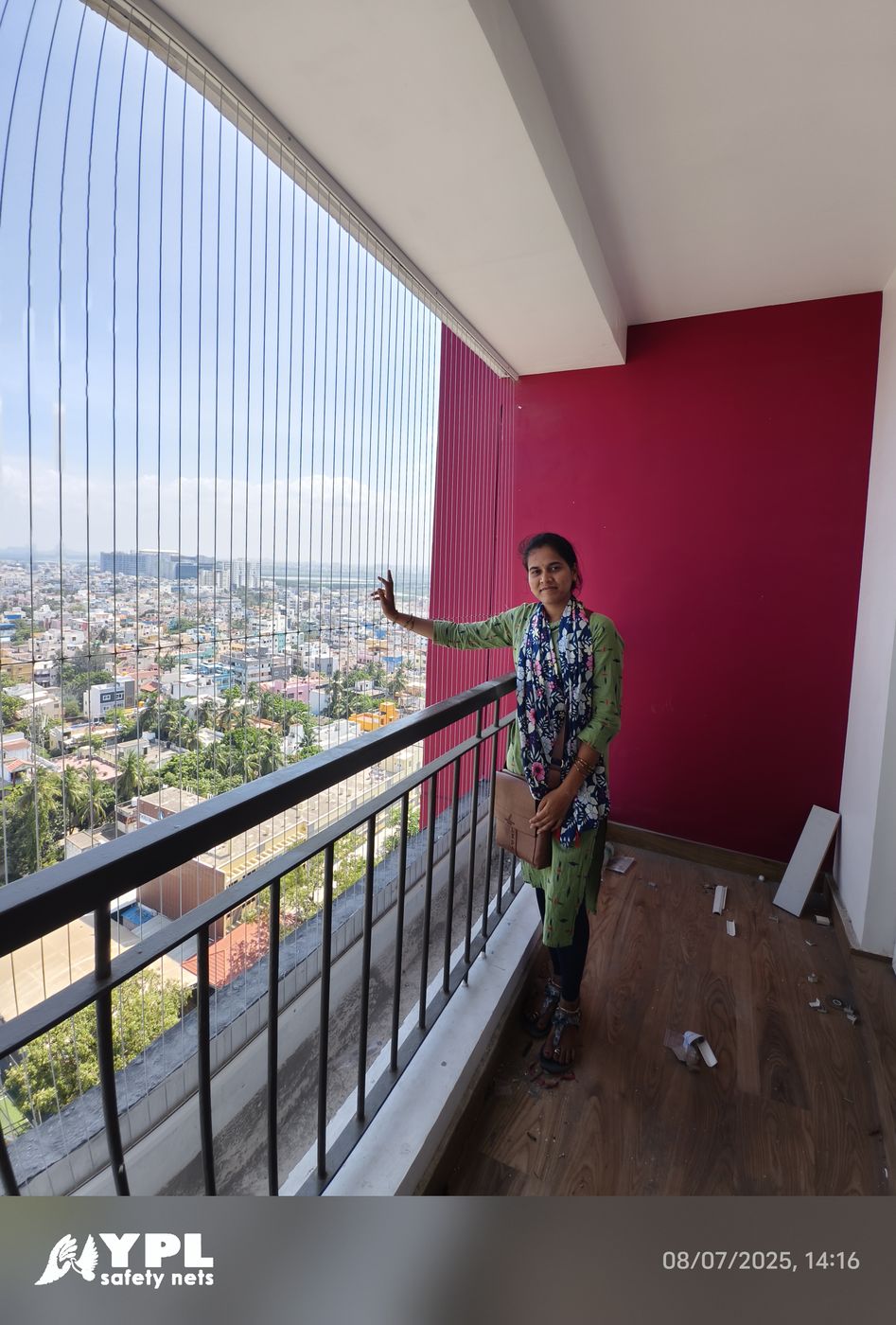 Woman beside vertical invisible grill wires on a high-rise balcony with a red wall and expansive city views.