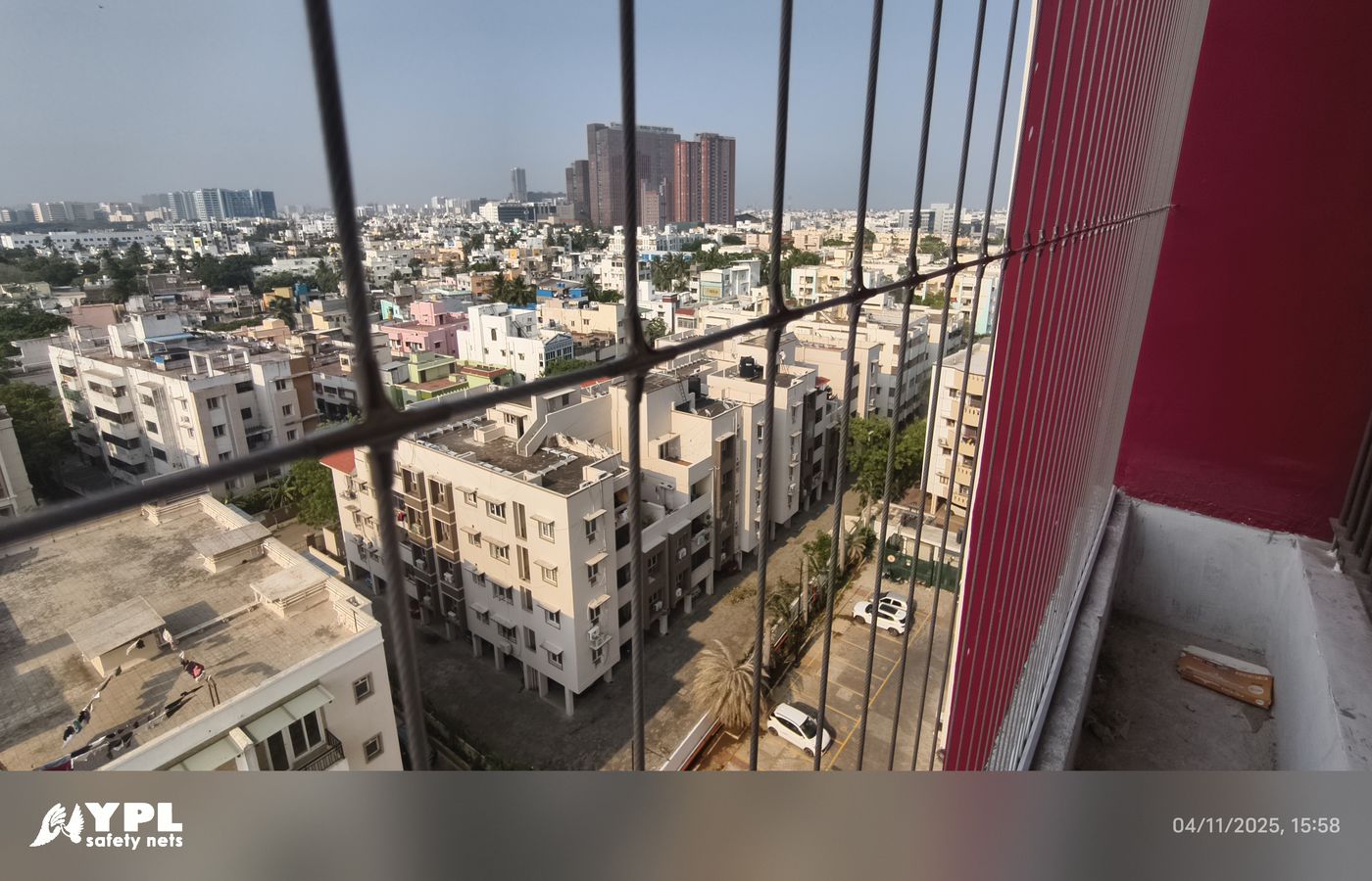 Close view through balcony grill wires overlooking apartment buildings and streets, with a red wall and narrow concrete ledge.