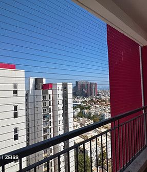 High-rise balcony with horizontal invisible grill wires, black railing, and red wall overlooking neighboring towers under blue skies.