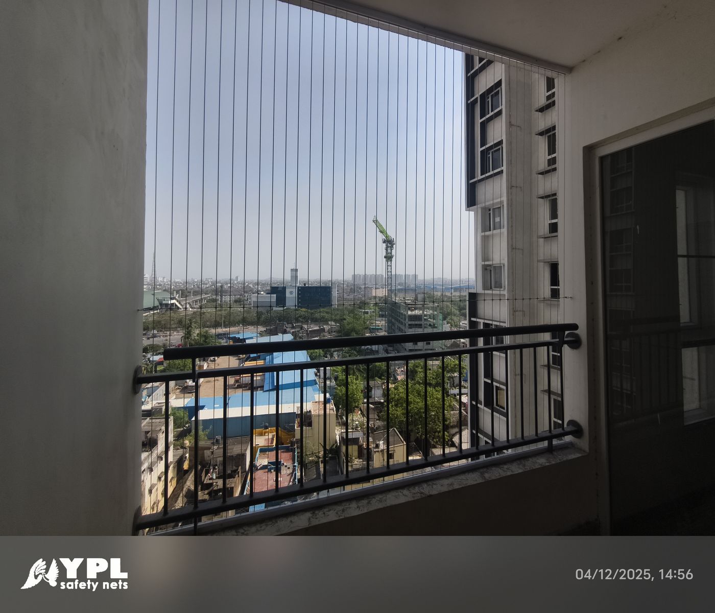 Apartment balcony fitted with vertical invisible grill wires above black railings, overlooking buildings and a construction crane.