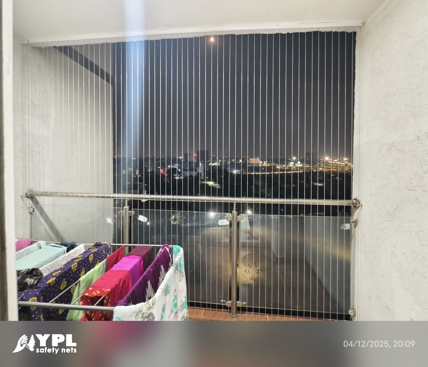 Balcony with vertical invisible grills, glass railing, and drying clothes overlooking city lights at night.