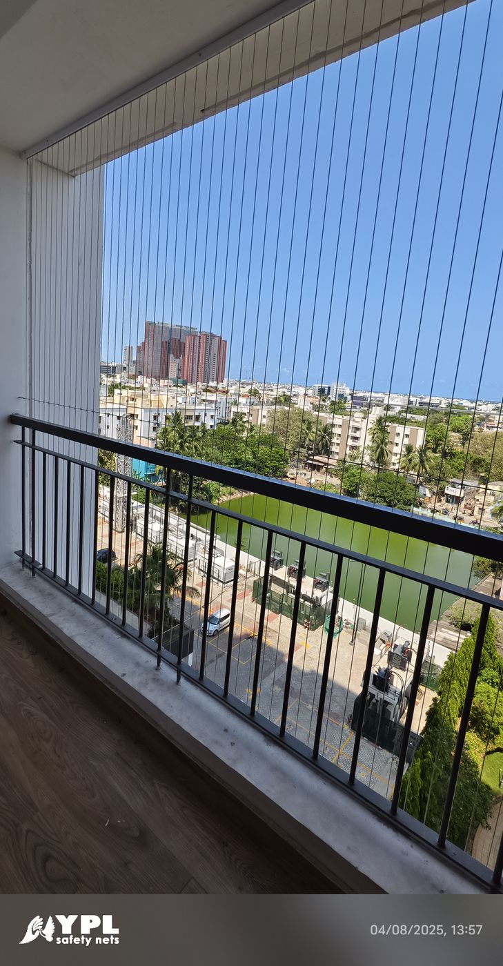 Sunny balcony with invisible grills, black metal railing, and wood-look flooring overlooking buildings and a green sports field.