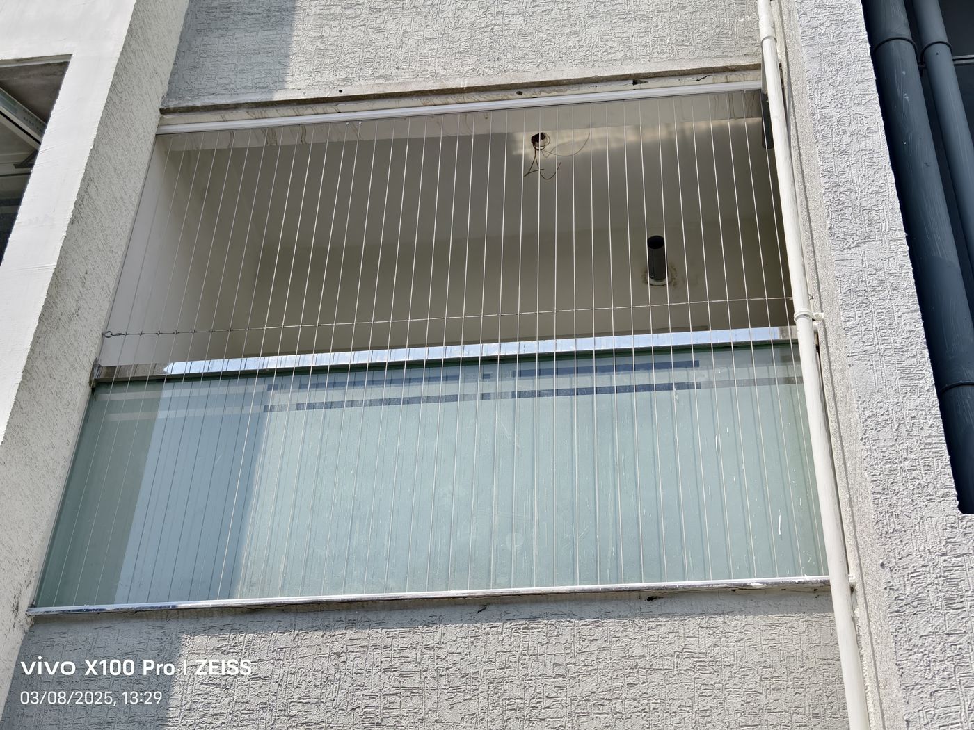 Exterior view of recessed balcony fitted with vertical invisible grills above frosted glass panels, beside exposed pipes.