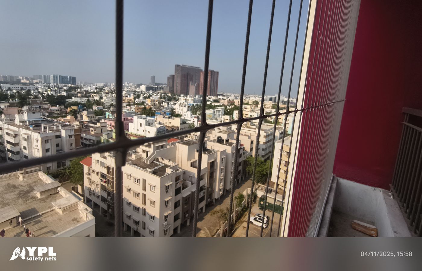 View through metal bars and fine vertical grill wires toward apartment buildings, beside a red balcony wall.