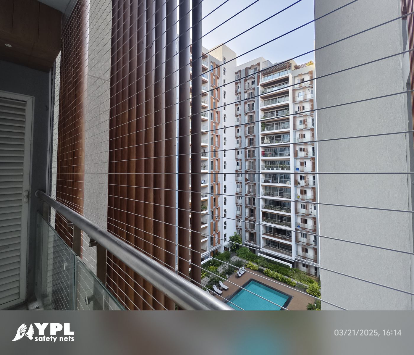 Horizontal invisible grill wires across a high-rise balcony overlooking apartment towers and a swimming pool.