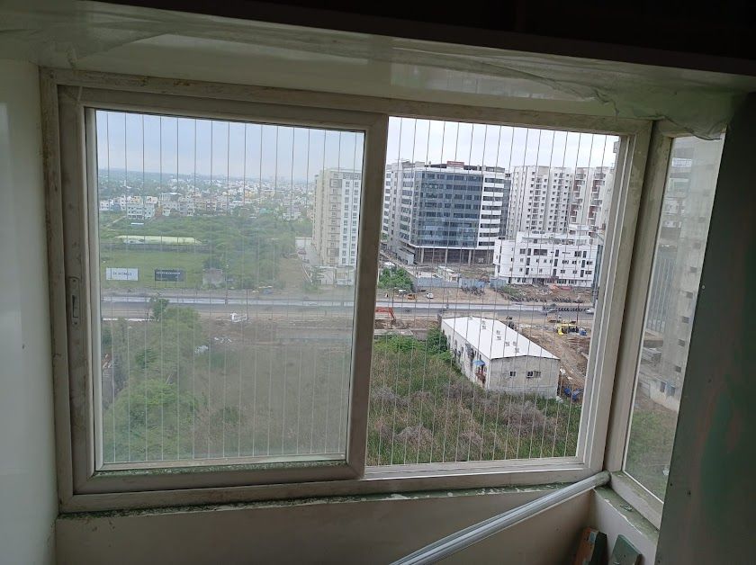 Interior view through sliding windows with vertical invisible grills overlooking roads, greenery, and high-rise buildings.