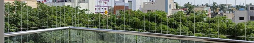 Cropped balcony view showing invisible grill wires above glass corner railings, with trees and residential buildings beyond.