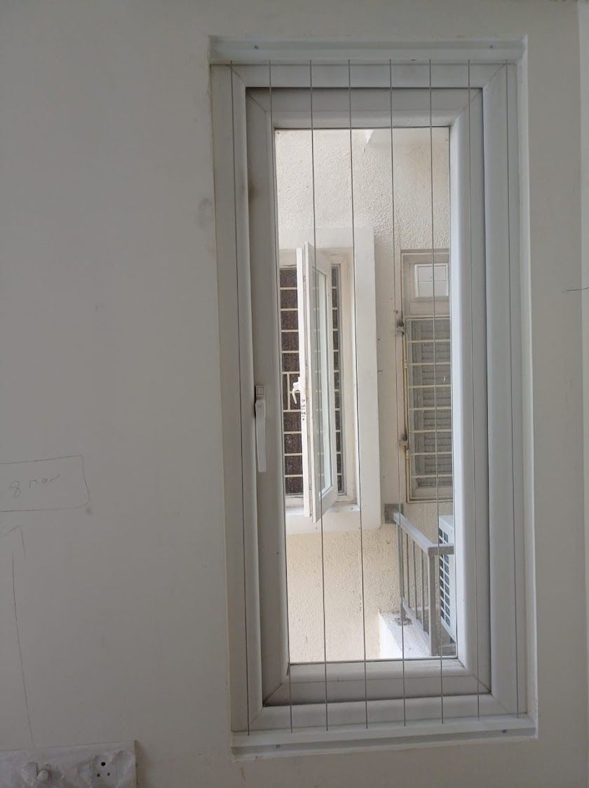 Narrow white-framed window with vertical invisible grills overlooking a building duct with windows and an air-conditioning unit.