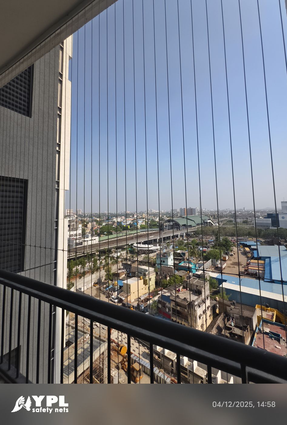 High-rise balcony with vertical invisible grill cables and black railing overlooking roads, rooftops, and an elevated transport corridor.