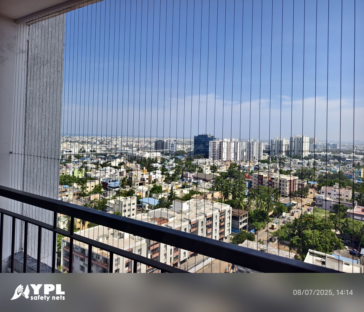 High-rise balcony with vertical invisible grill cables overlooking a dense cityscape under blue skies.