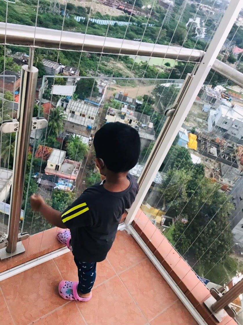 Child standing on a tiled high-rise balcony enclosed by vertical safety wires, overlooking buildings and greenery.