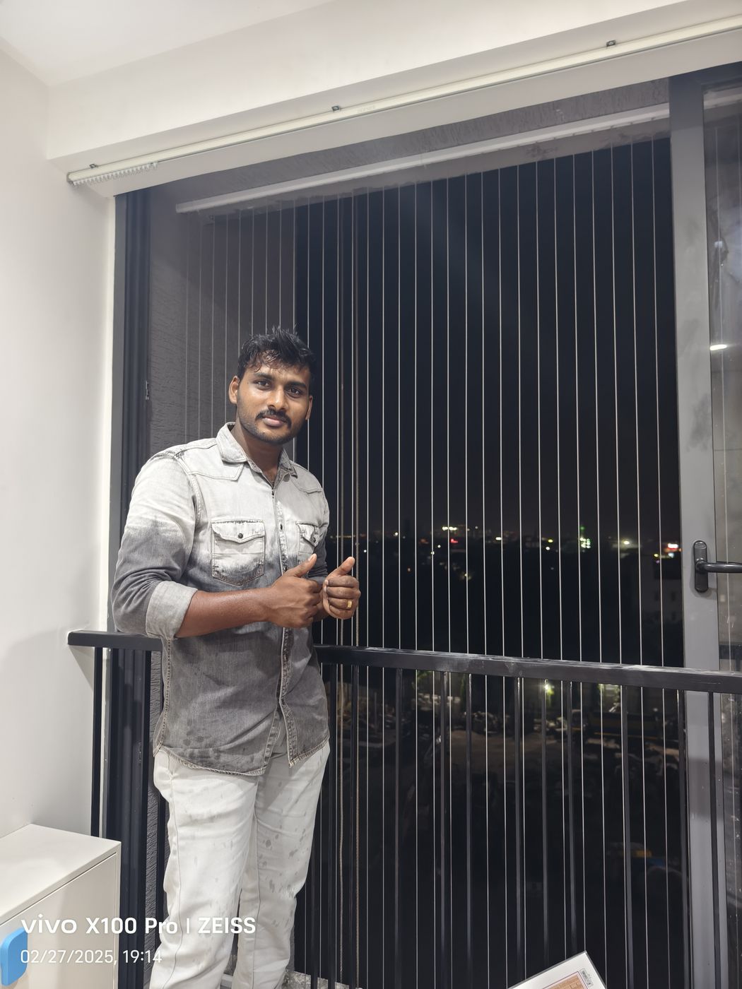 Man giving thumbs up beside vertical invisible grills and a black balcony railing, with city lights visible at night.
