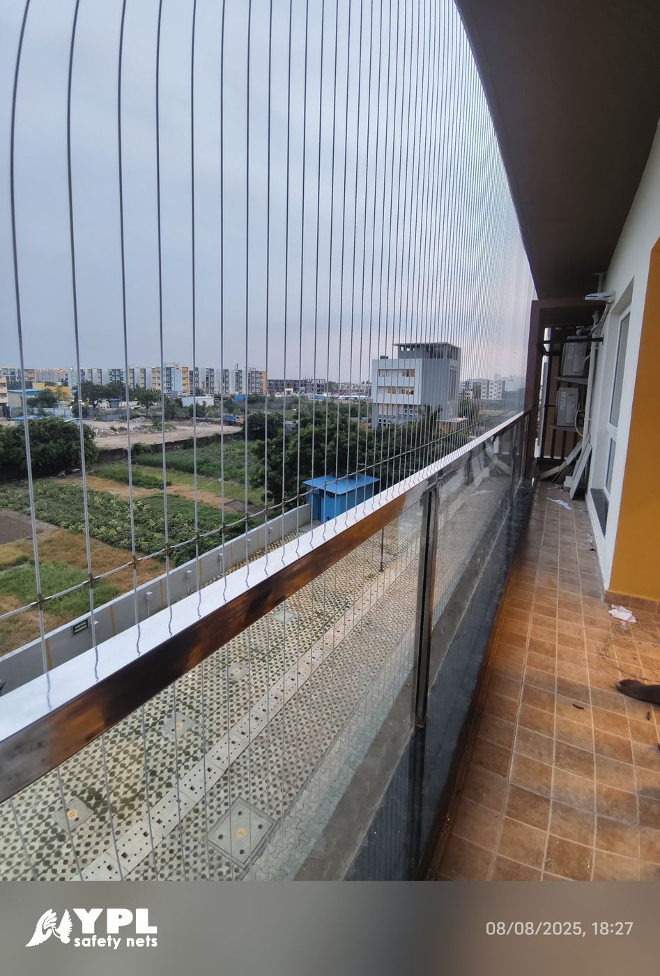 Long tiled apartment balcony with vertical invisible grills and glass railing overlooking green plots and city buildings.