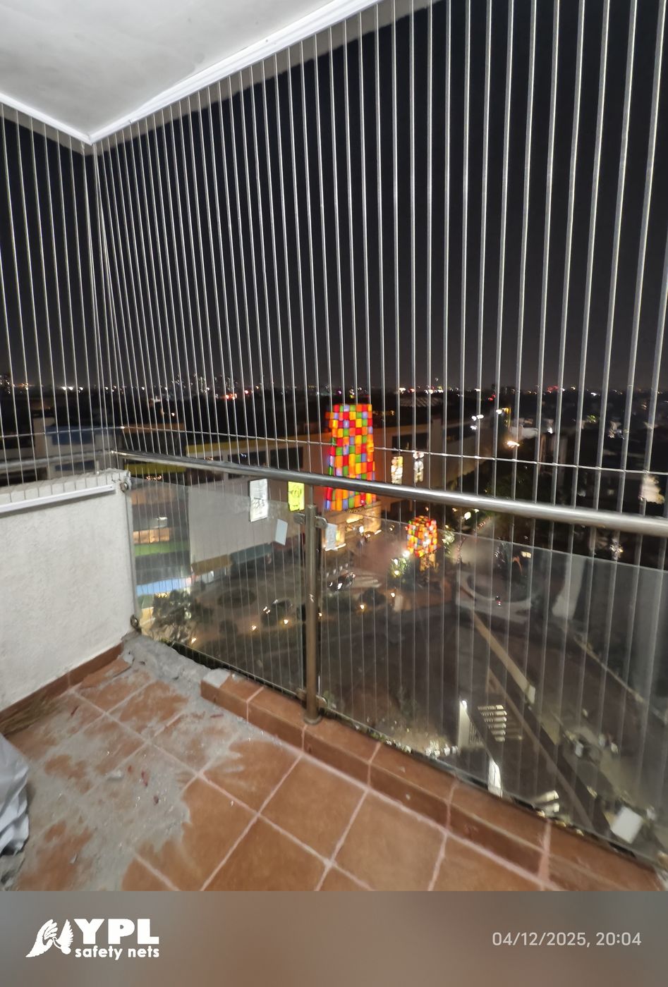 Corner balcony enclosed with vertical invisible grills above glass railings, overlooking brightly lit buildings and streets at night.