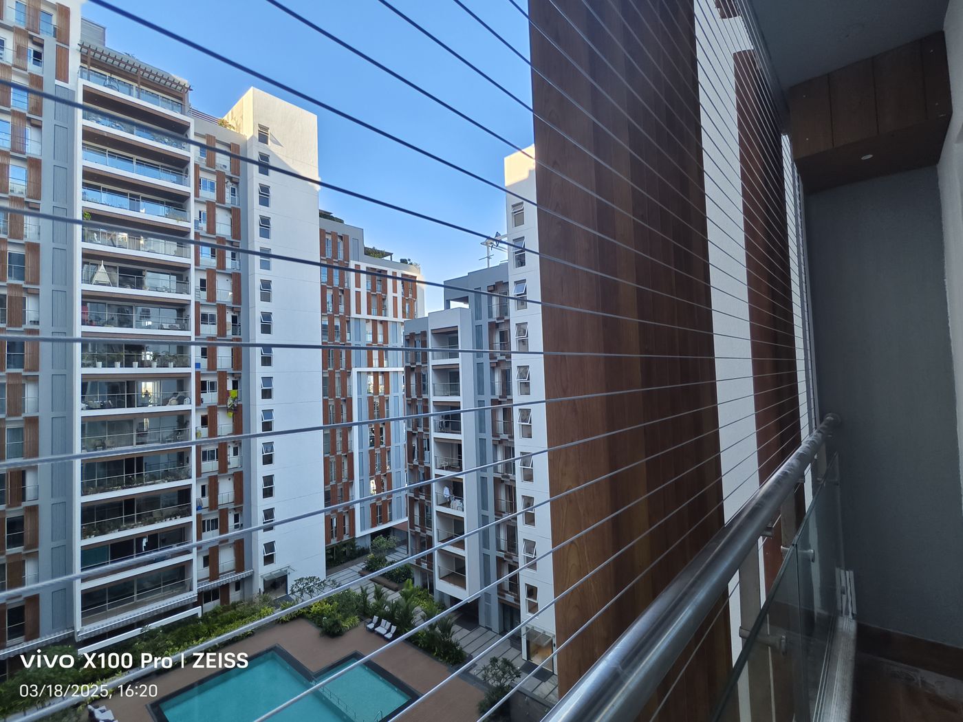 Horizontal invisible grills enclose a high-rise balcony overlooking apartment towers and a swimming pool.