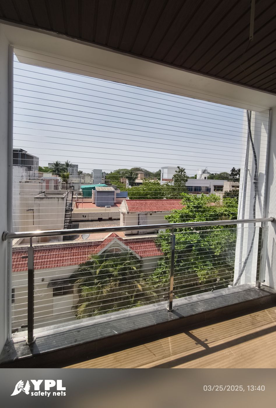 Sunlit balcony with horizontal invisible grills, metal railing, and views of low-rise homes and leafy trees.