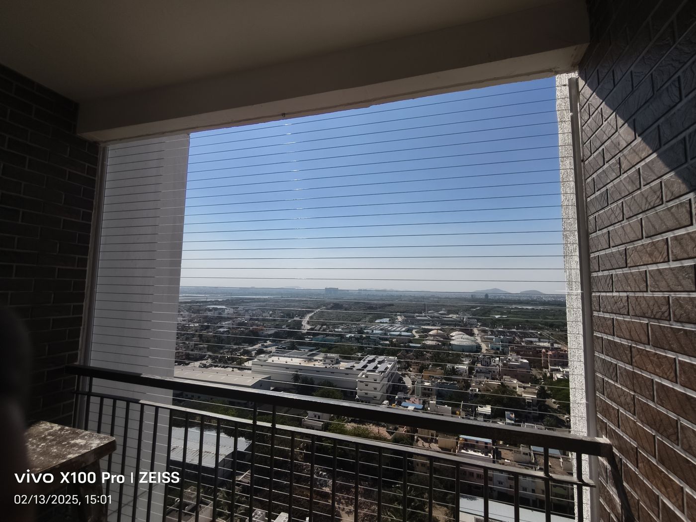 Brick-lined high-rise balcony with horizontal invisible grills and a black railing overlooking an expansive cityscape.