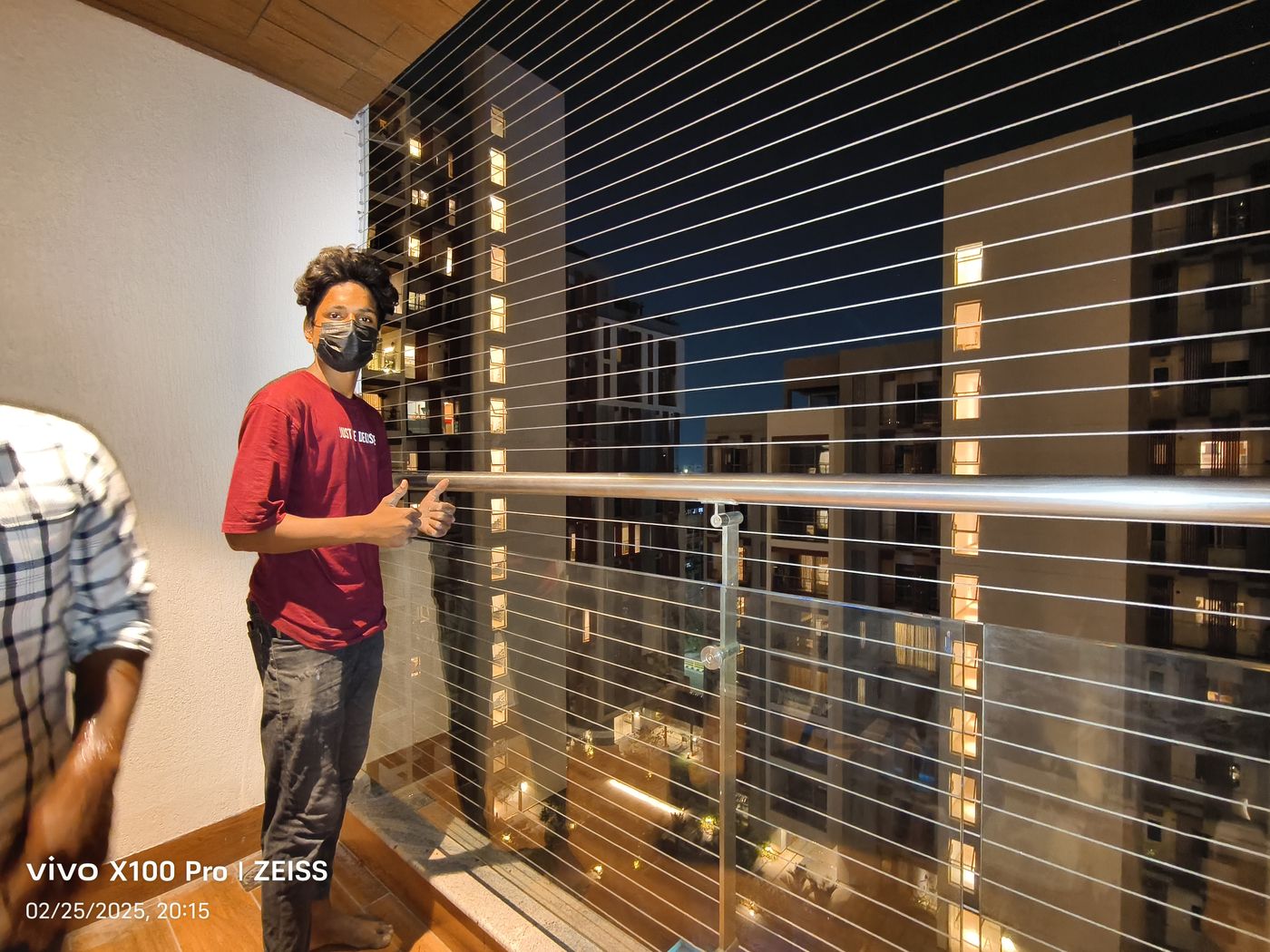 Masked person beside horizontal invisible grills on a balcony overlooking illuminated apartment towers at night.