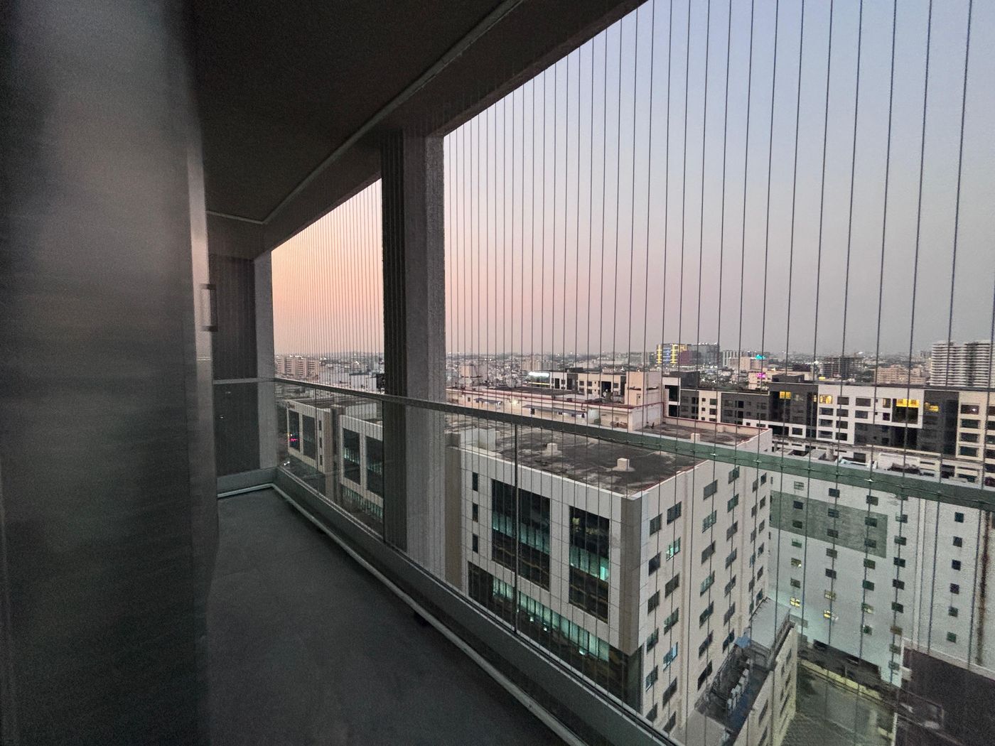 Long high-rise balcony with vertical invisible grills and glass railing overlooking urban buildings at dusk.