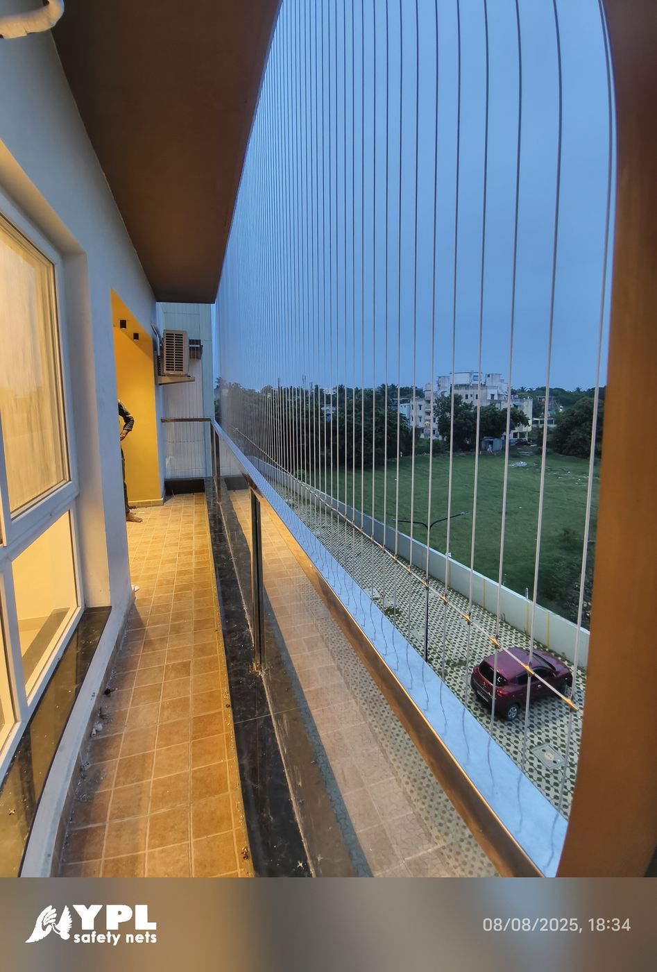Vertical invisible grills enclose a narrow tiled balcony overlooking a green field and nearby buildings at dusk.