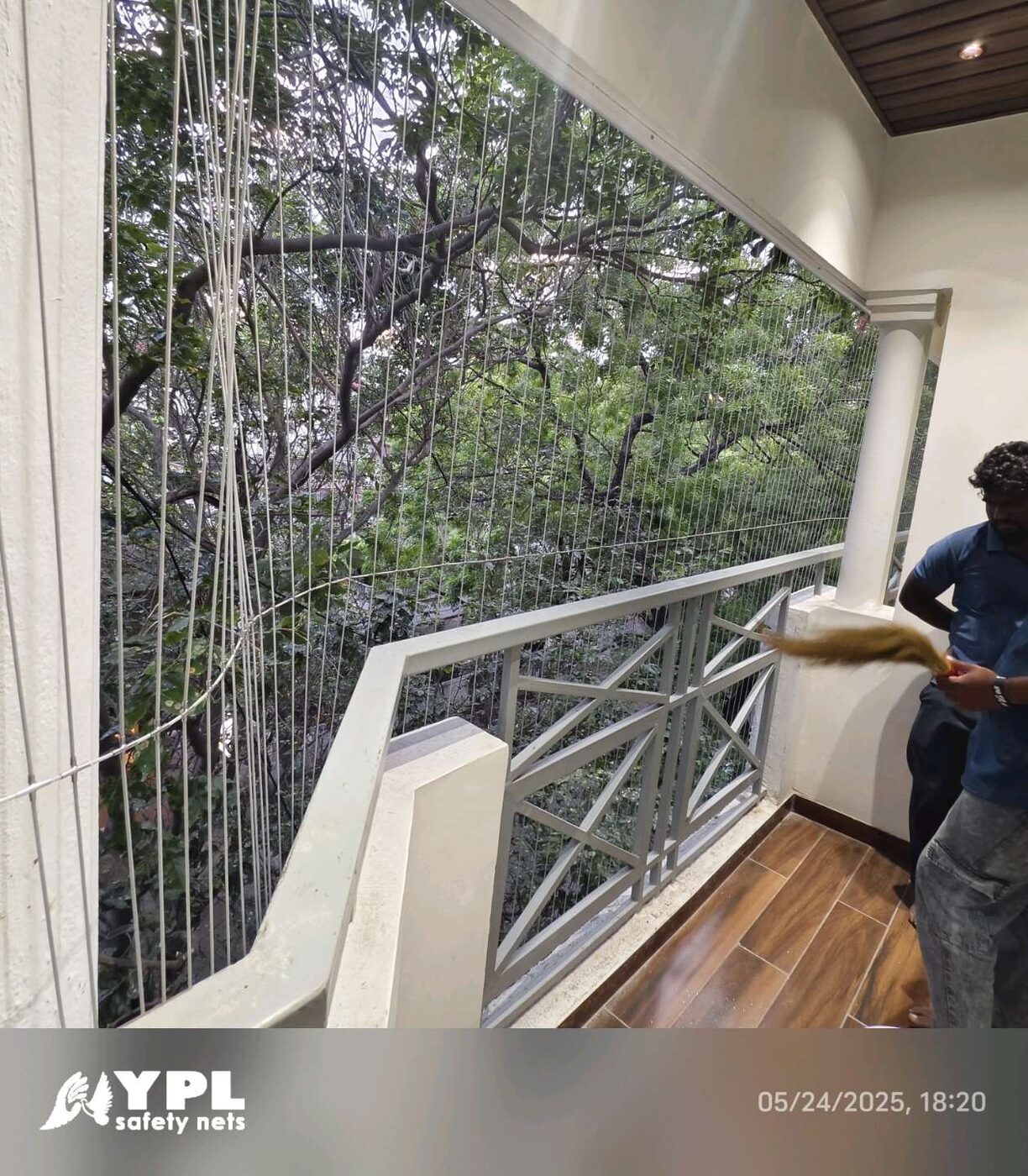Vertical invisible grills line a tiled balcony with geometric metal railings, leafy trees outside, and workers at one side.