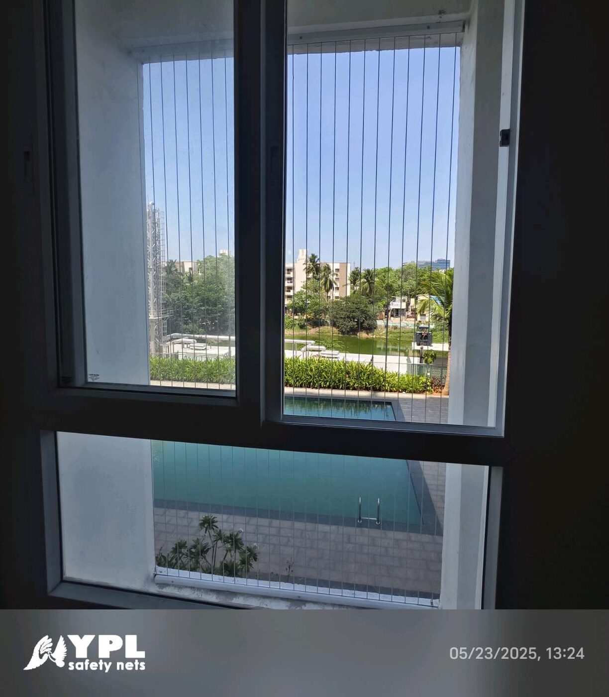 Interior view through a sliding window with vertical invisible grills, overlooking a swimming pool, landscaped grounds, and buildings.