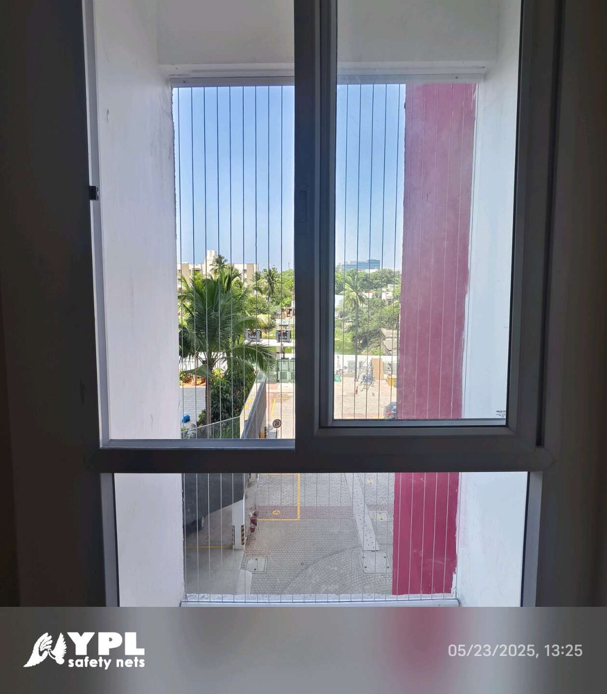 Sliding window fitted with vertical invisible grills overlooks palm trees and a paved courtyard beside a red exterior column.