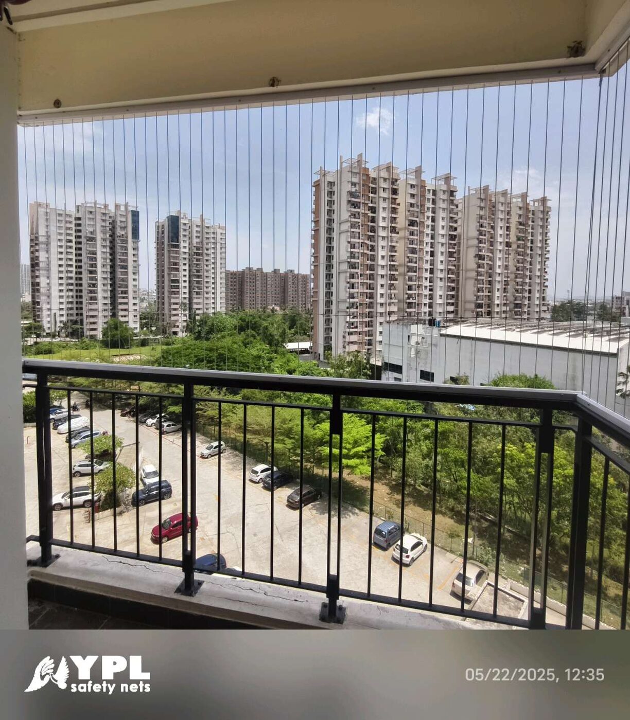Balcony with vertical invisible grills overlooking apartment towers, trees, and a parking area in daylight.