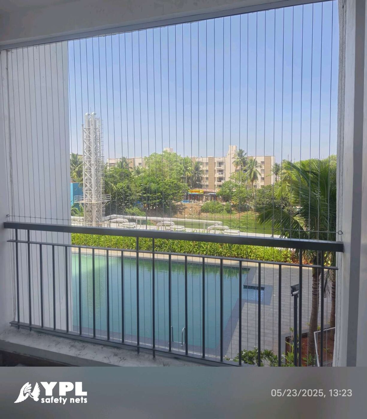 Balcony with vertical invisible grills and black railing overlooking a swimming pool, palm trees, and residential buildings.