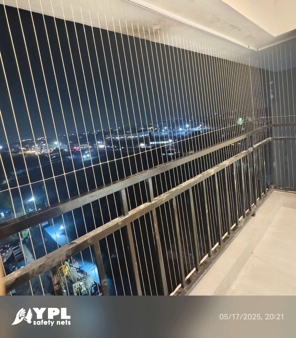 Balcony enclosed with vertical invisible grills above metal railings, overlooking city lights at night.