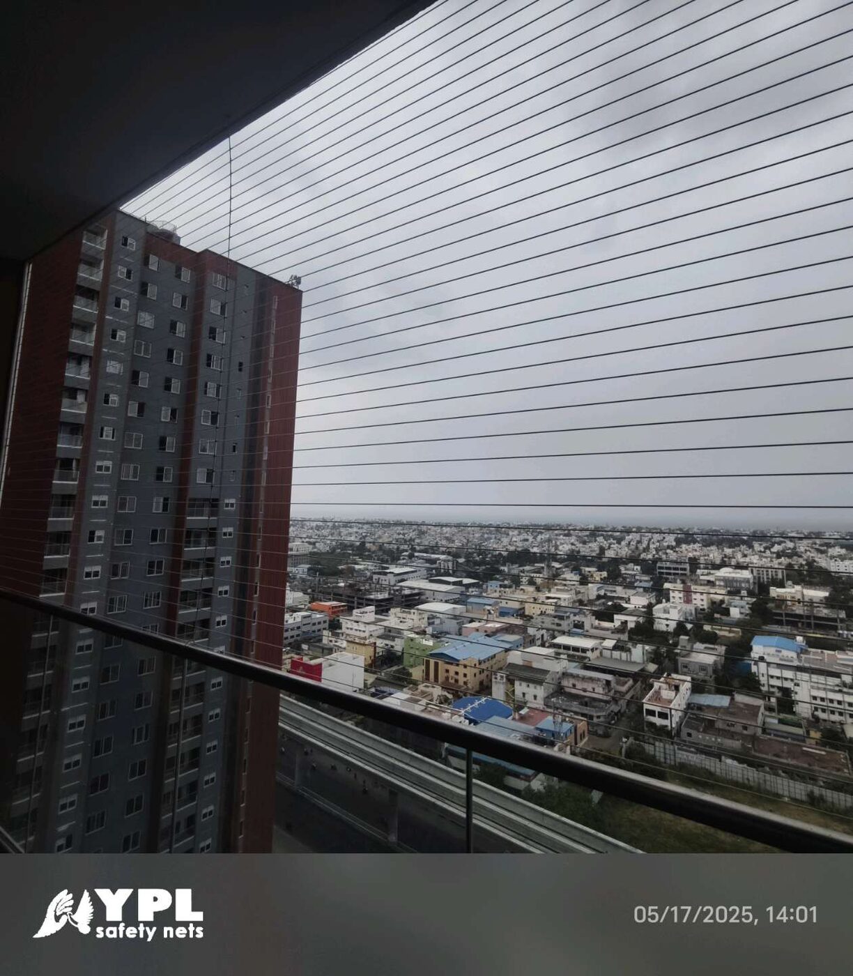 High-rise balcony with horizontal invisible grills and glass railing overlooking a neighboring tower and sprawling cityscape.
