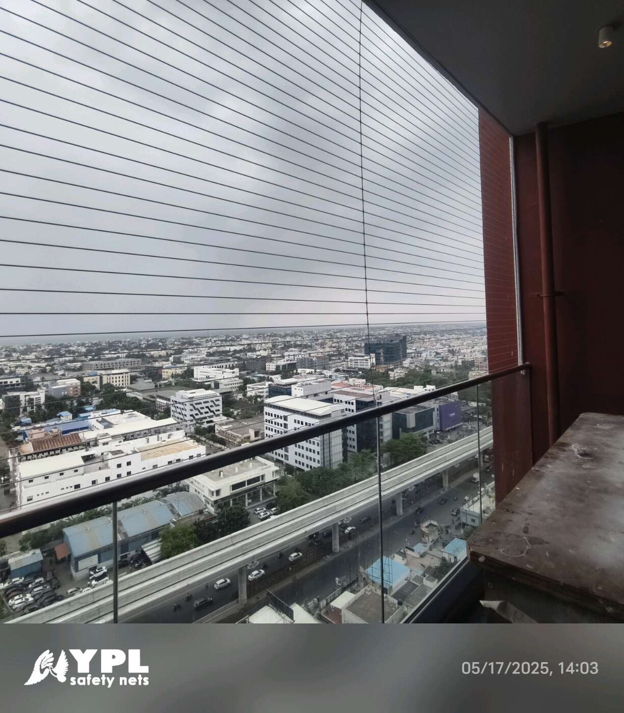 Horizontal invisible grills enclose a high-rise balcony with glass railing, overlooking city buildings and an elevated roadway.