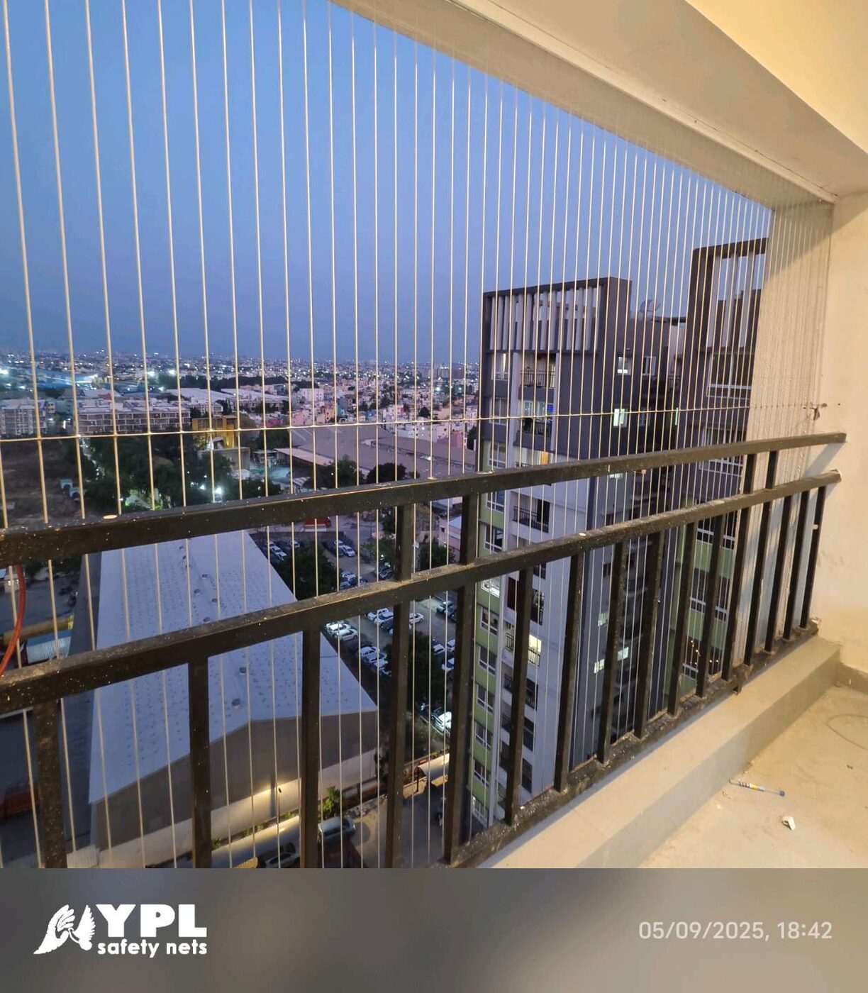 Vertical invisible grills above a black balcony railing, overlooking neighboring apartment towers and city lights at dusk.