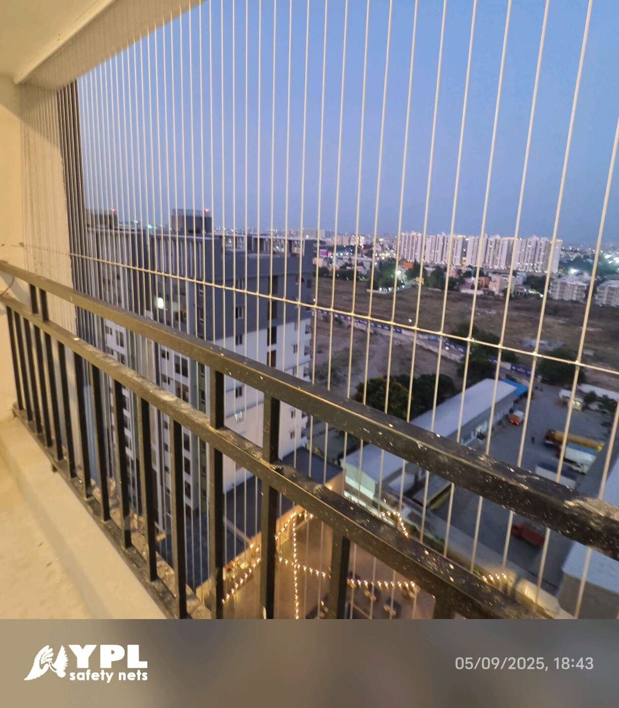 High-rise balcony with vertical invisible grills and black railing, overlooking apartment buildings and open land at dusk.