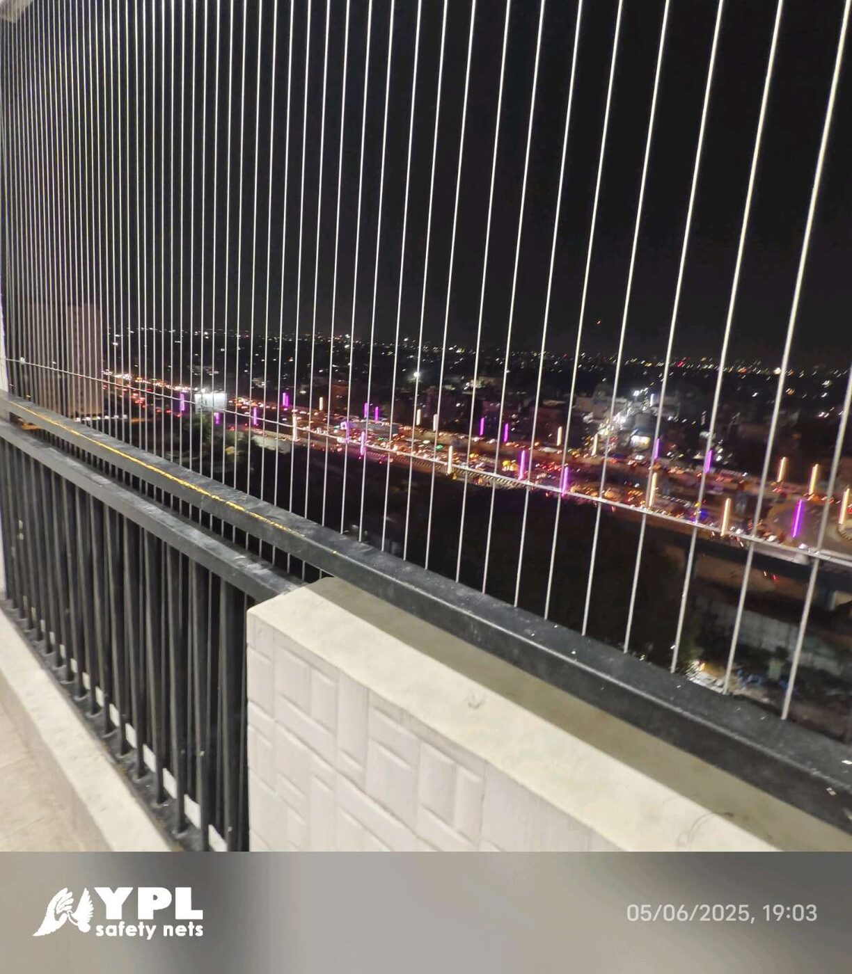Vertical invisible grills line a high-rise balcony with black railing, overlooking a brightly illuminated road at night.