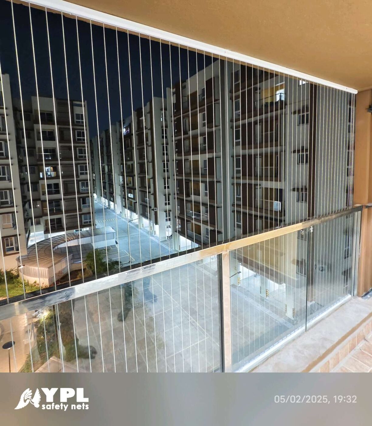 Balcony with vertical invisible grills and glass railing overlooking illuminated apartment buildings at night.