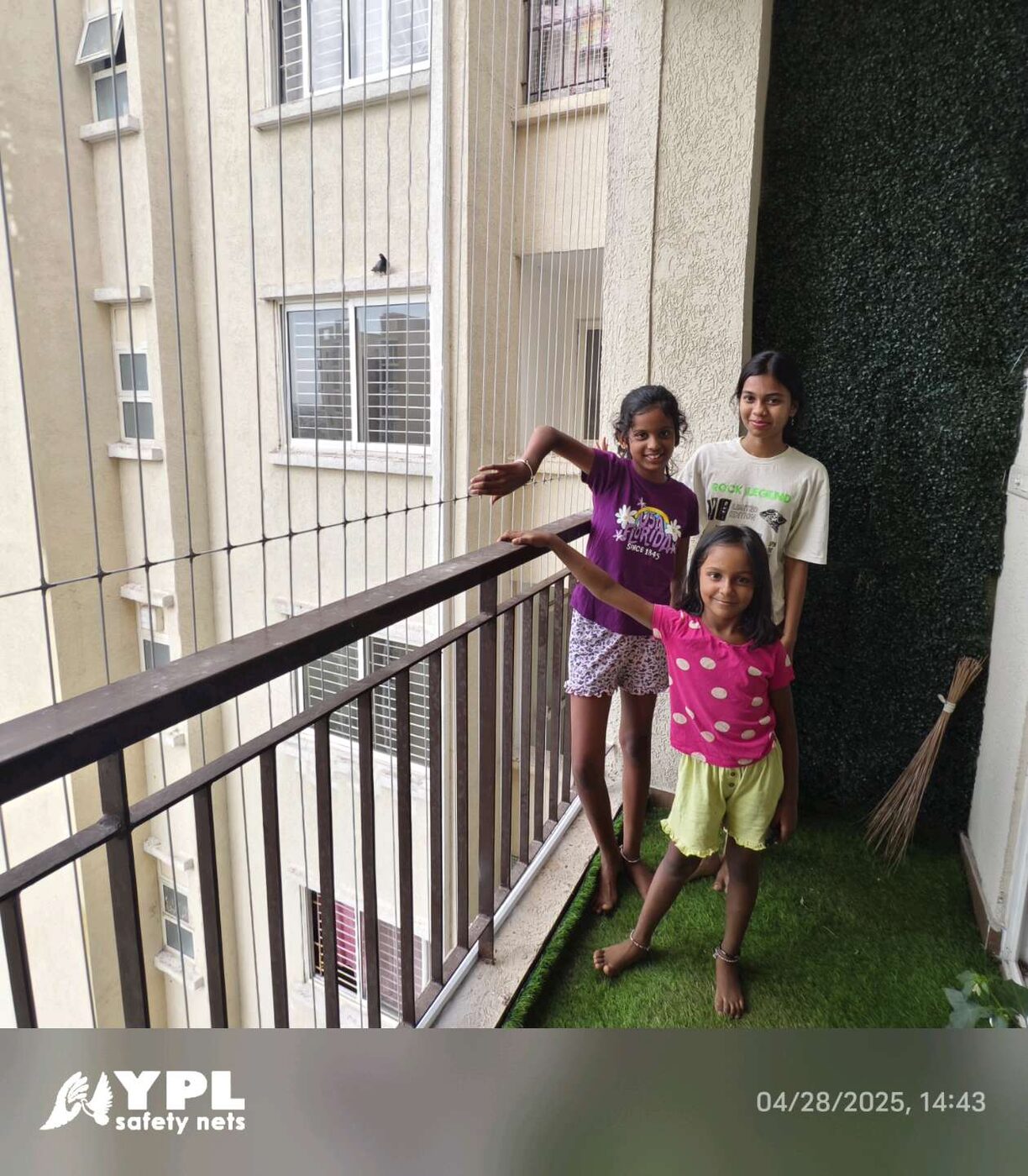 Three children on a narrow apartment balcony with invisible grills, artificial grass flooring, and a green accent wall.