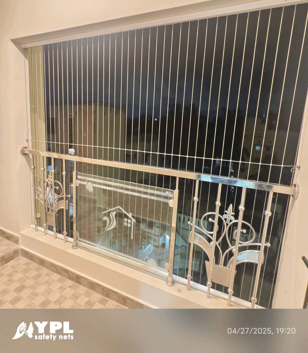Vertical invisible grills above a decorative metal and glass balcony railing, viewed from a tiled interior at night.