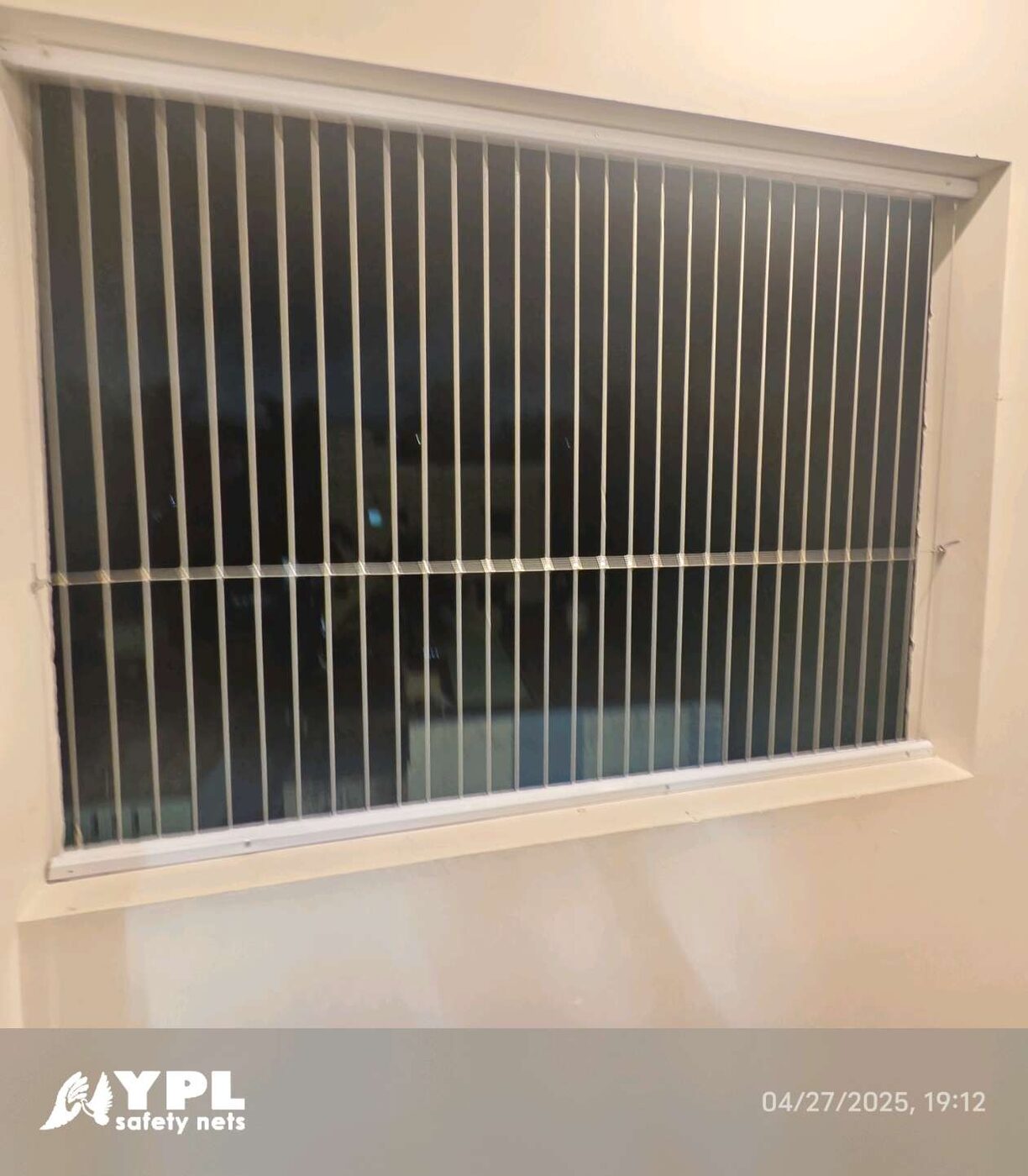Rectangular window opening fitted with vertical invisible grills and a horizontal support wire, viewed indoors at night.
