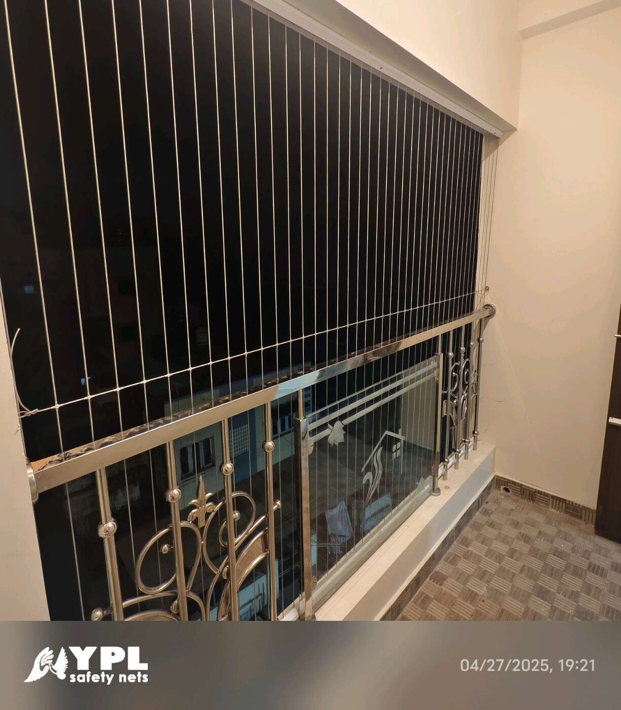 Vertical invisible grills above a decorative metal balcony railing, with tiled flooring and neighboring buildings visible at night.
