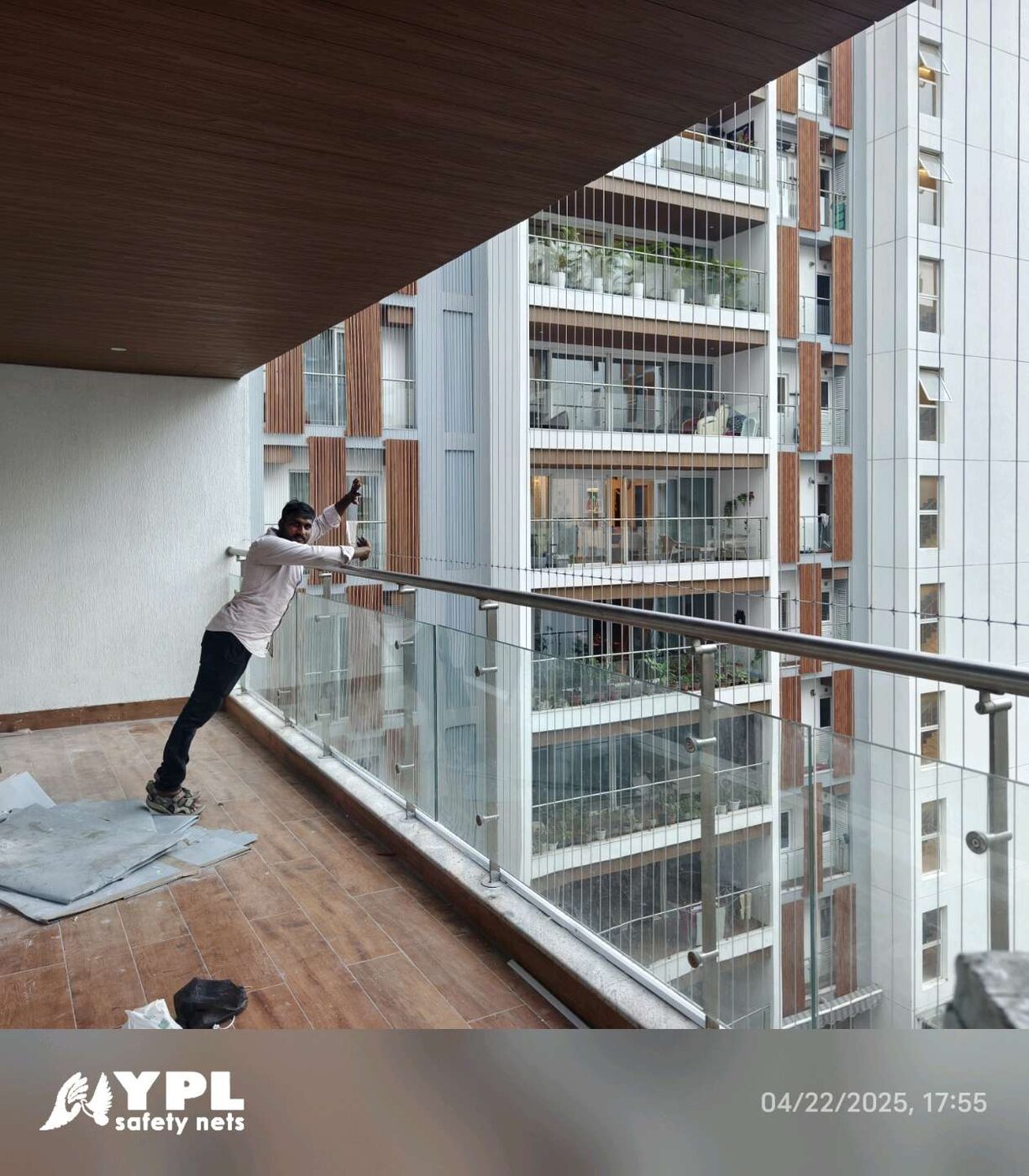 Worker fitting invisible grills on a high-rise balcony with glass railings, wood-look flooring, and neighboring apartment views.