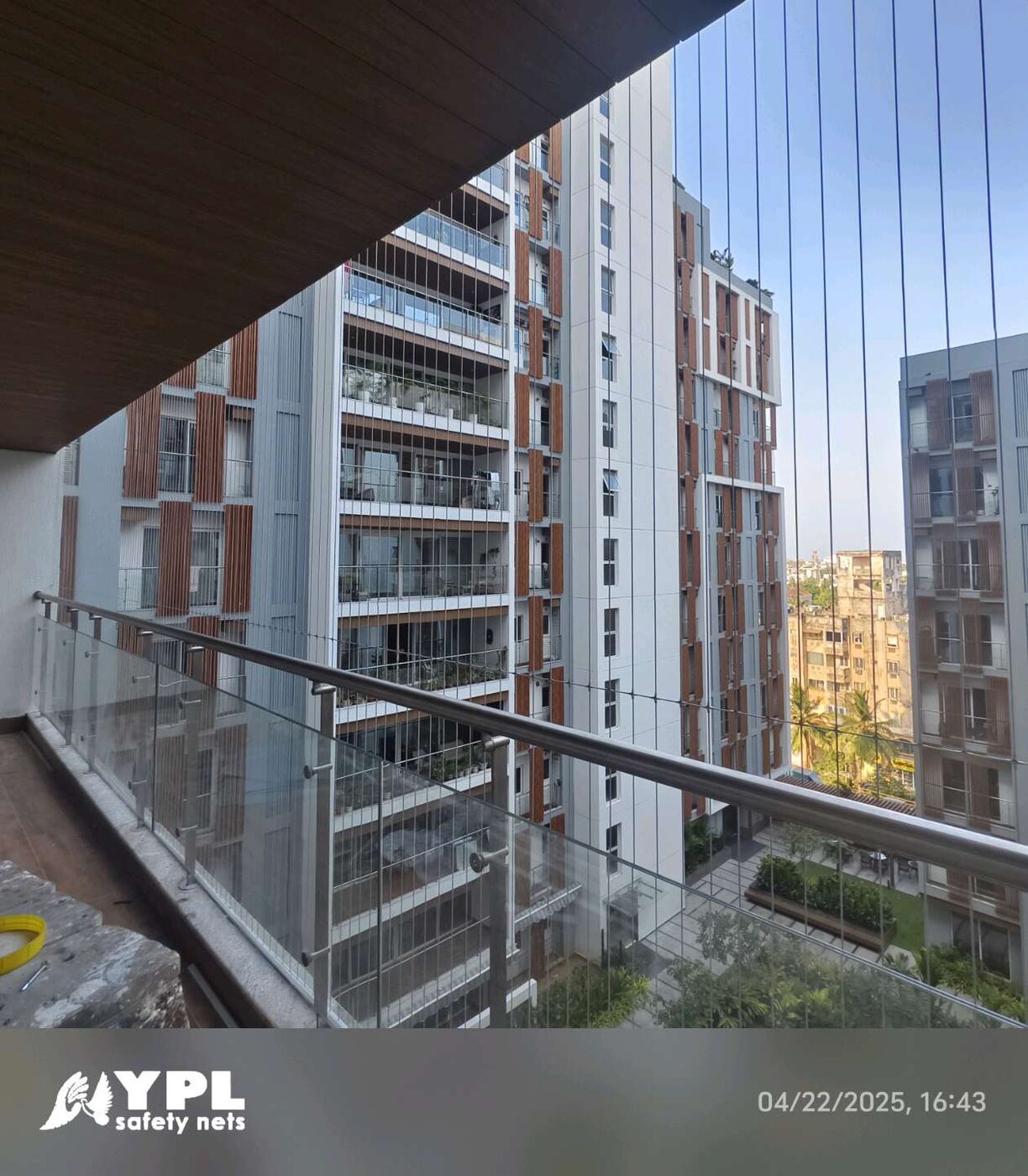 Vertical invisible grills enclose a glass-railed high-rise balcony overlooking apartment towers and landscaped grounds.