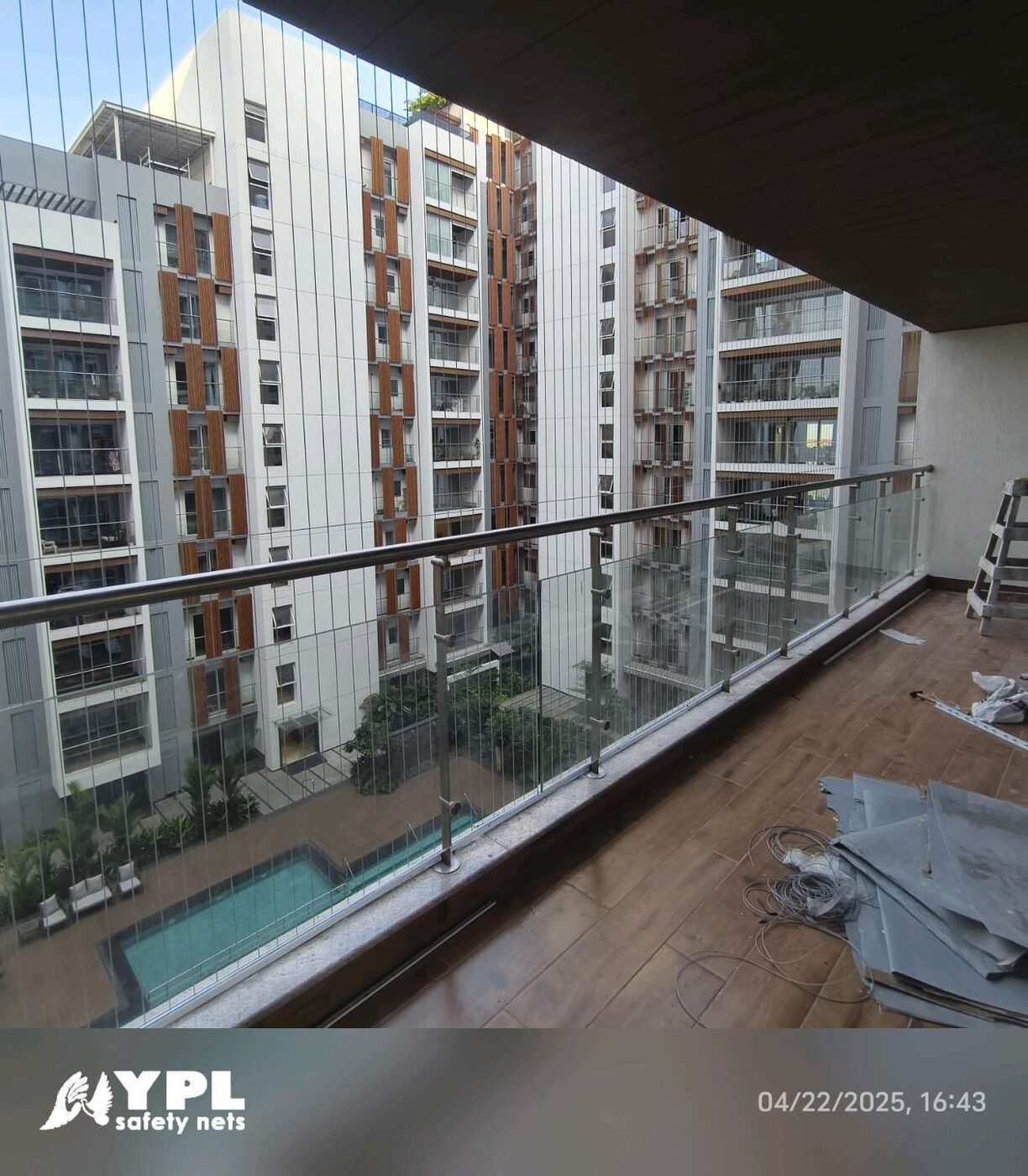 Invisible grills span a glass-railed balcony overlooking apartment towers and a swimming pool, with installation materials on the floor.