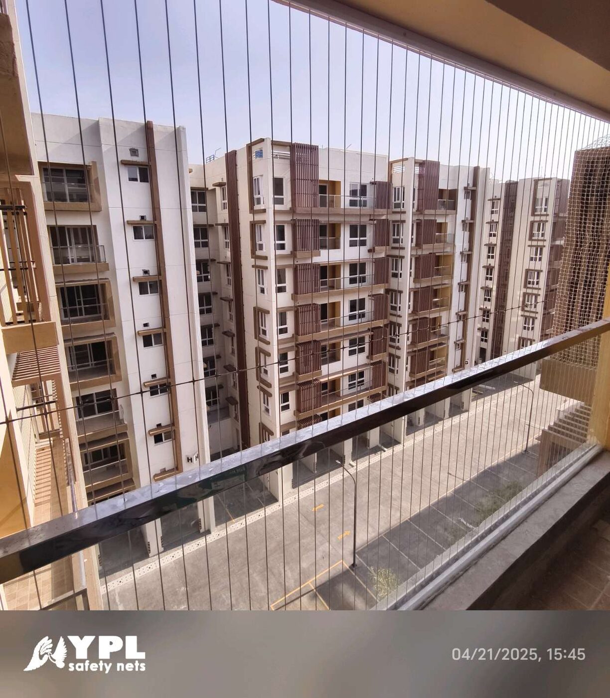 Vertical invisible grills line a covered balcony with a dark handrail, overlooking neighboring apartment blocks in daylight.