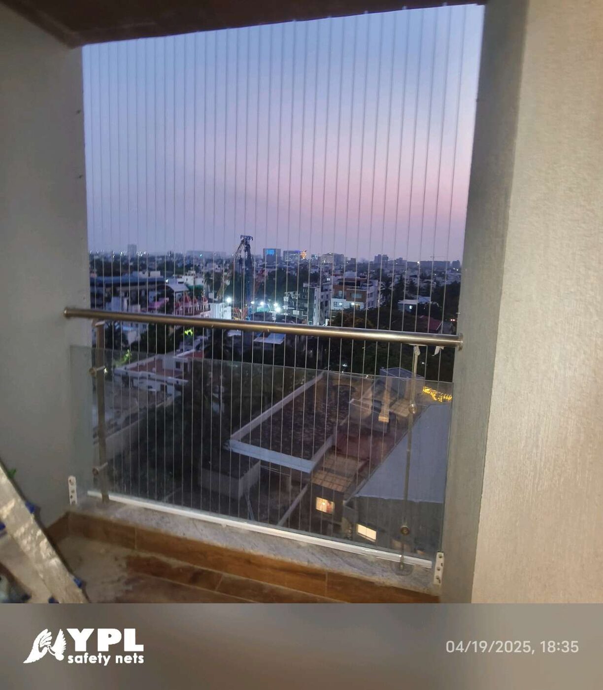 Balcony with vertical invisible grills and glass railing overlooking city rooftops at dusk, with a ladder nearby.