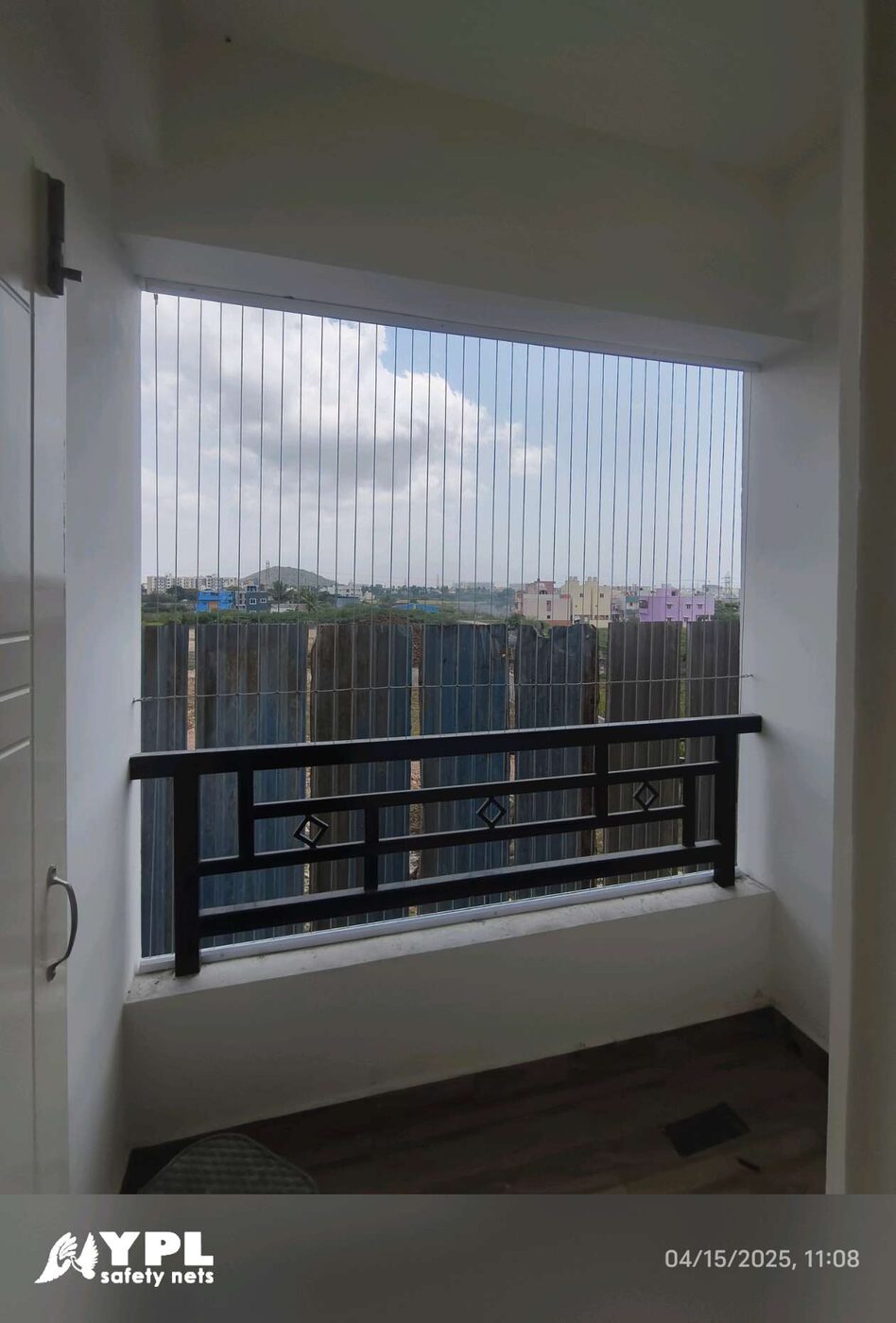 Recessed balcony opening with vertical invisible grills above a black railing, overlooking buildings and greenery under cloudy skies.