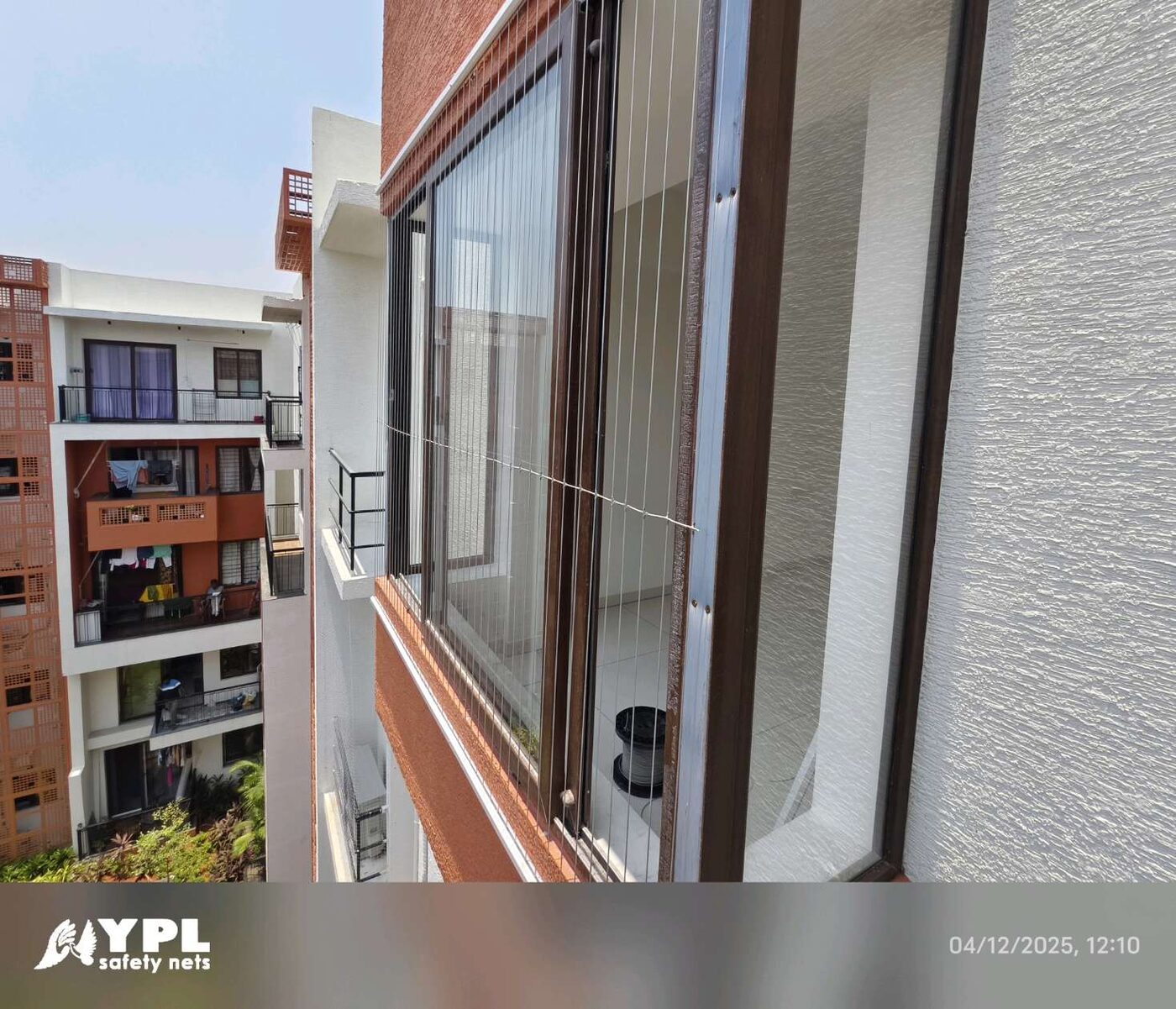 Angled exterior view of invisible grills across an apartment window, with neighboring balconies and courtyard greenery visible.