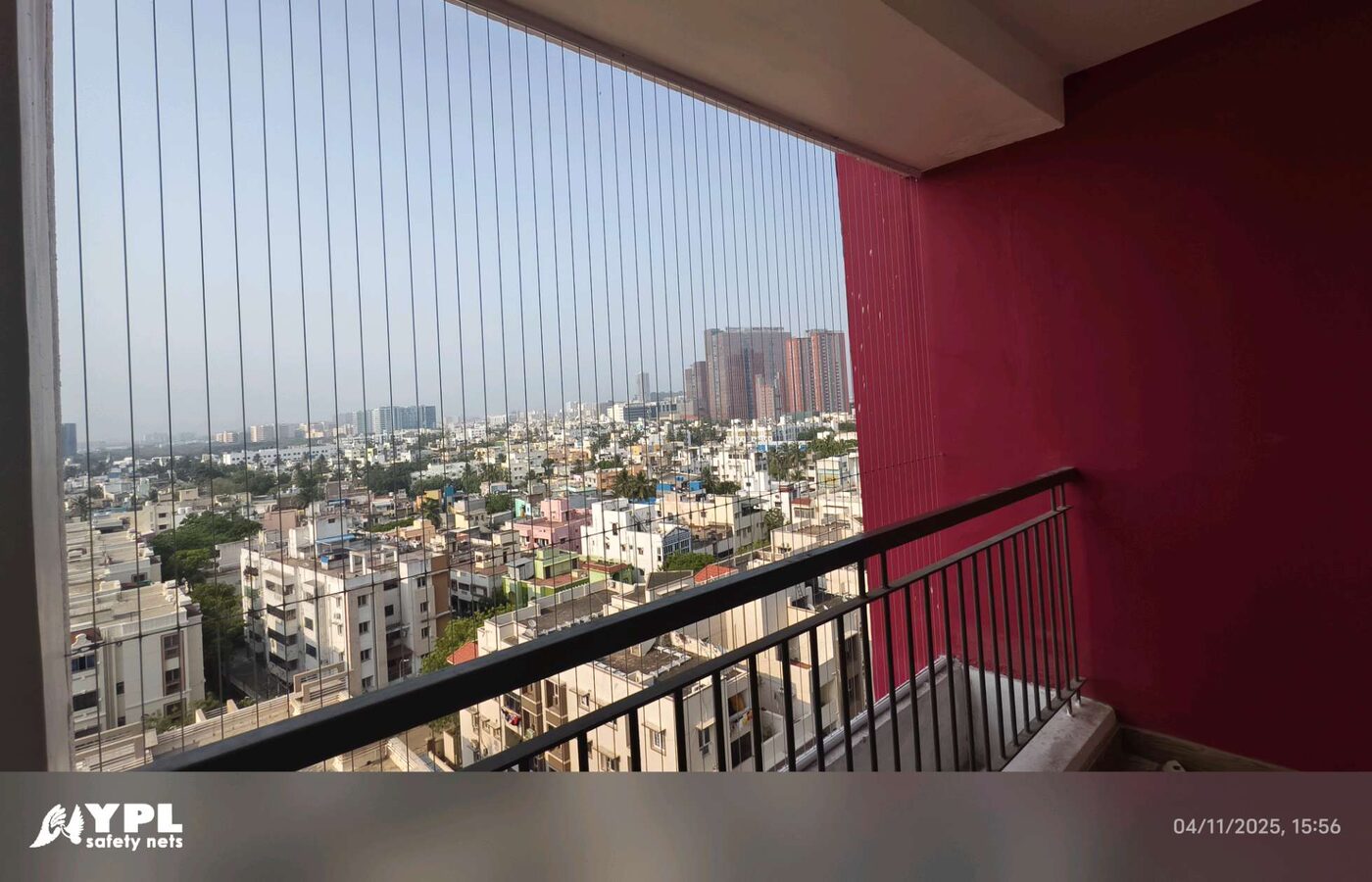 High-rise balcony with vertical invisible grill wires, black railing, and red wall overlooking dense city buildings.