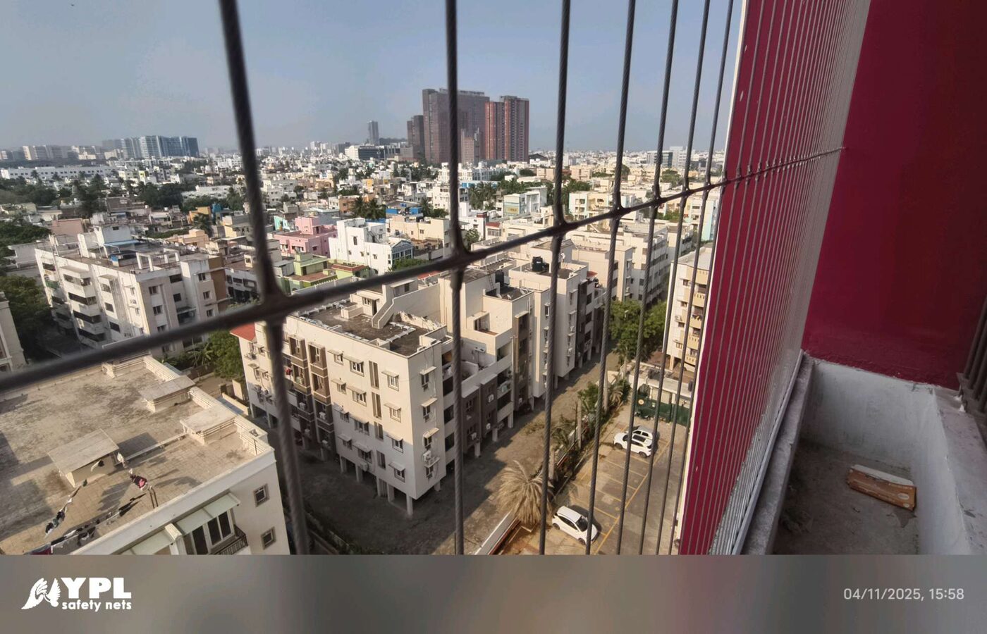Close view through balcony railing and invisible grill wires toward apartment buildings and a street below.